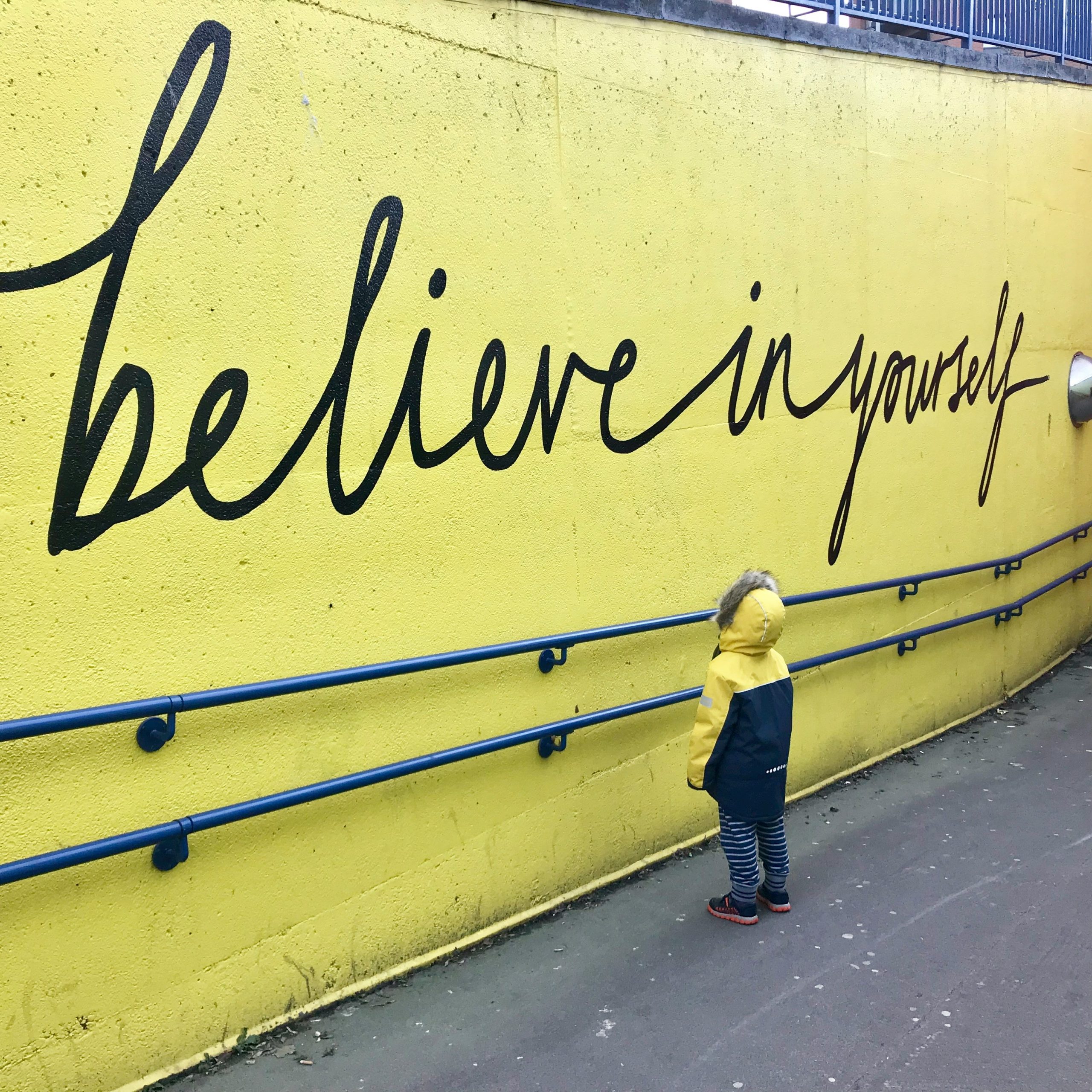 Kid standing in front of yellow wall that says, "believe in yourself".