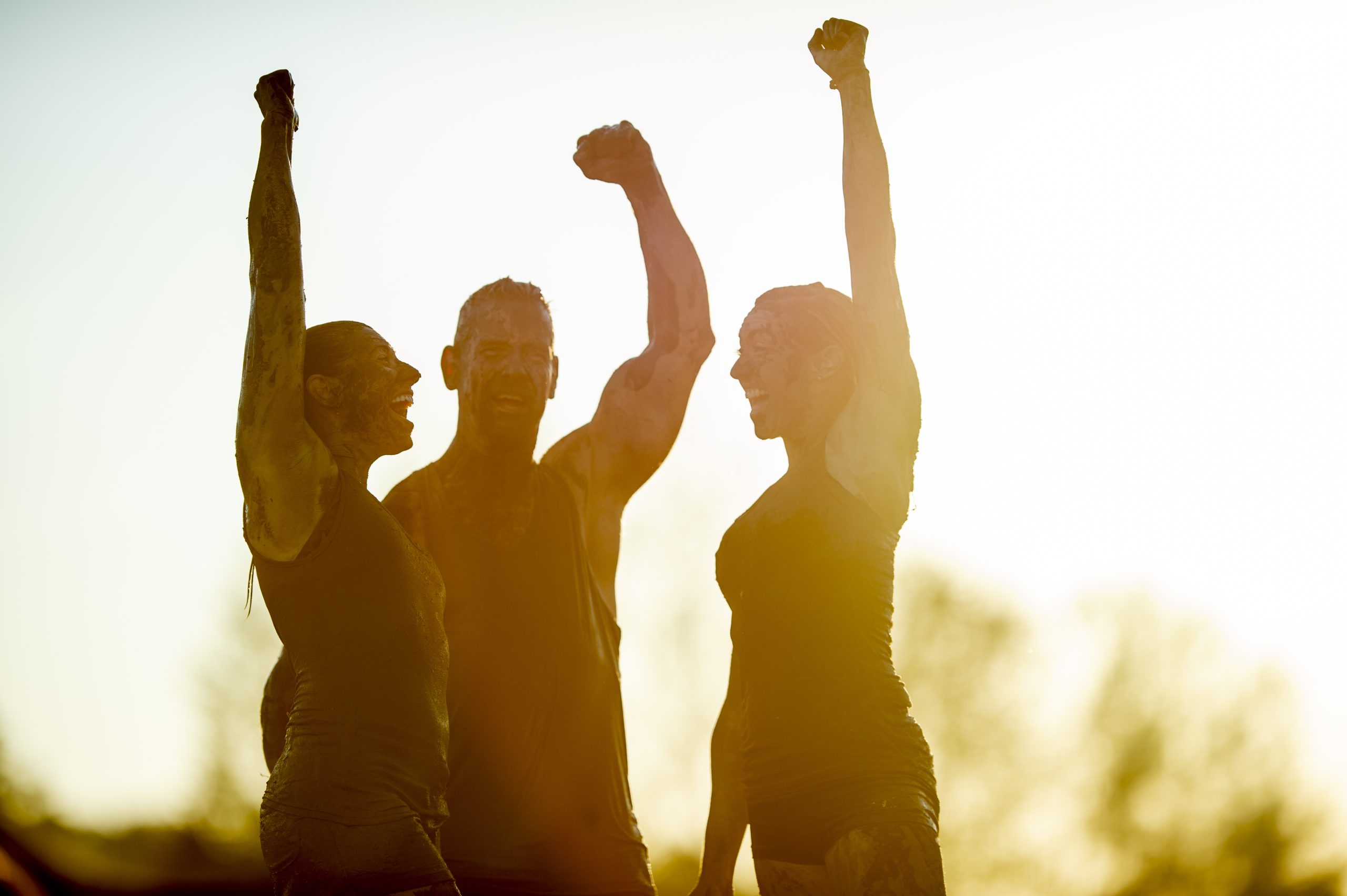 Three adult competitors stand side-by-side with their hands raised in the air as they cheer after a successful Mud Run win. They are each dressed comfortably in athletic wear and are covered in mud from head to toe.