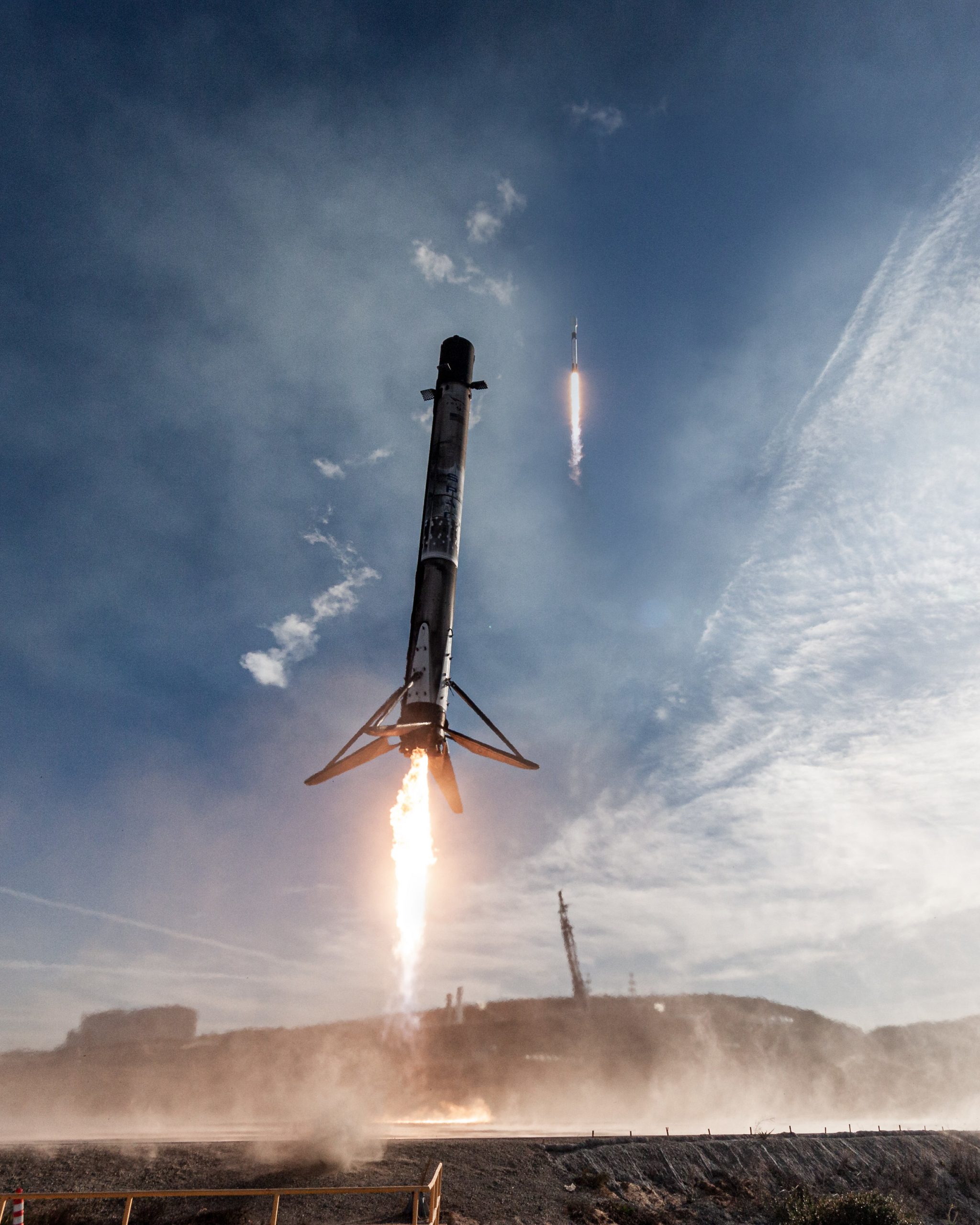 Photograph of the first stage of a Falcon 9 rocket returning to the ground as the rest of the rocket continues to space in the background.