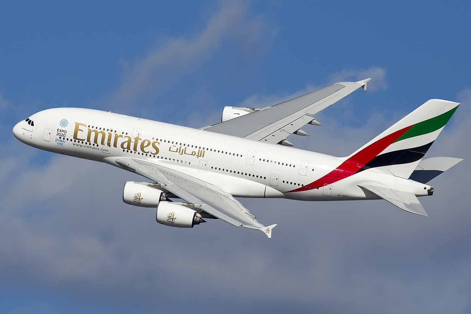 A white jet engine airplane flying towards the upper left corner with a partially cloudy background. Emirates is written on the fuselage.