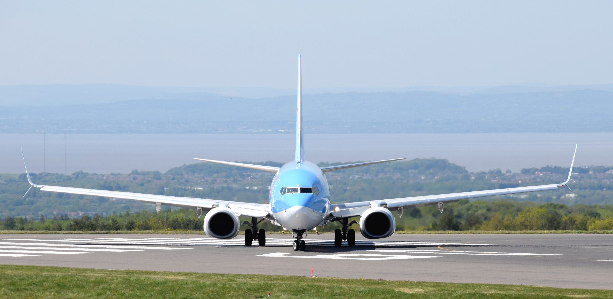A blue and white Boeing 737-800 aircraft facing the camera, moving down the airport runway. Water and low mountains in the background.