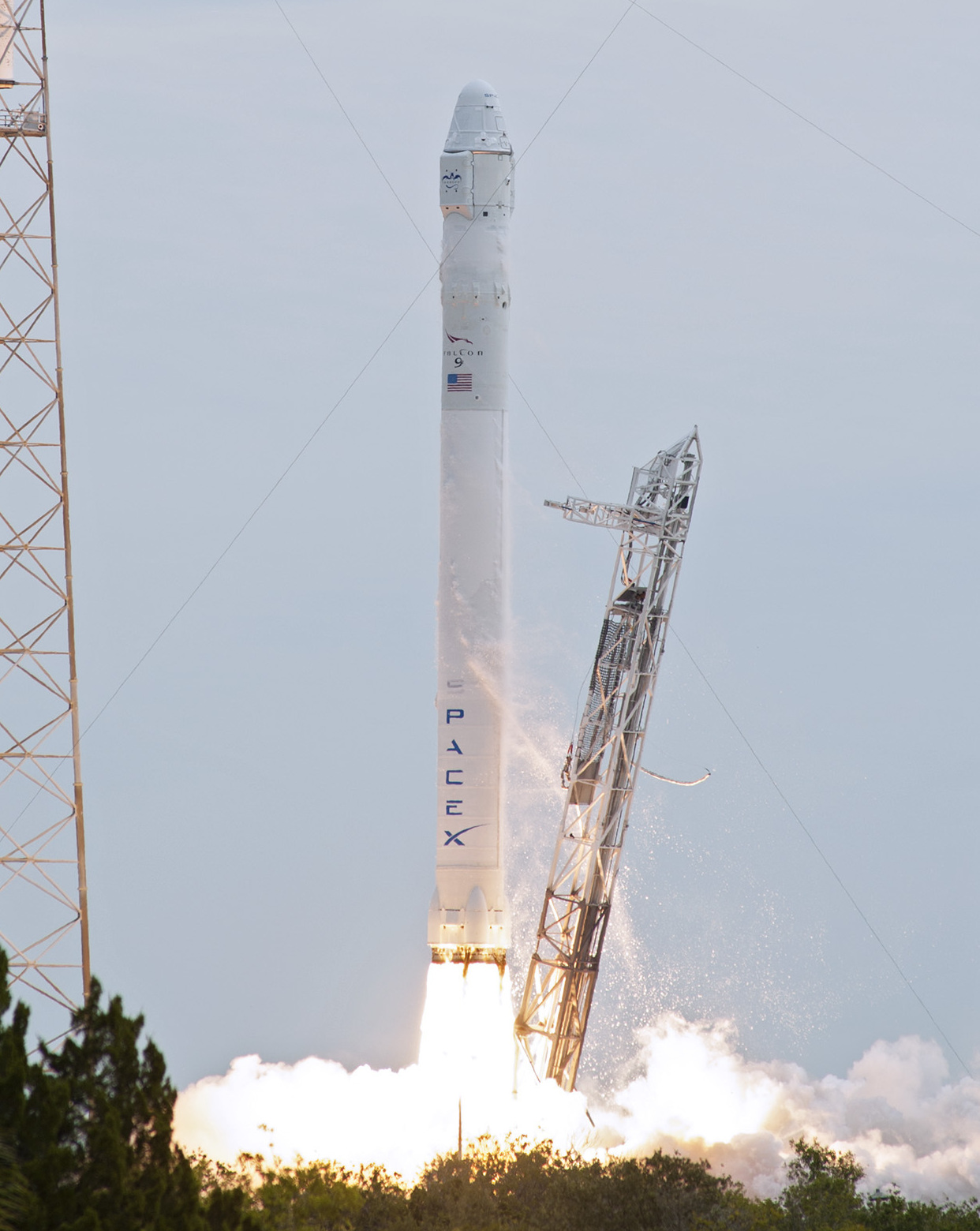 A large white rocket with SpaceX written on the side lifts off the platform while the frame falls away from it.