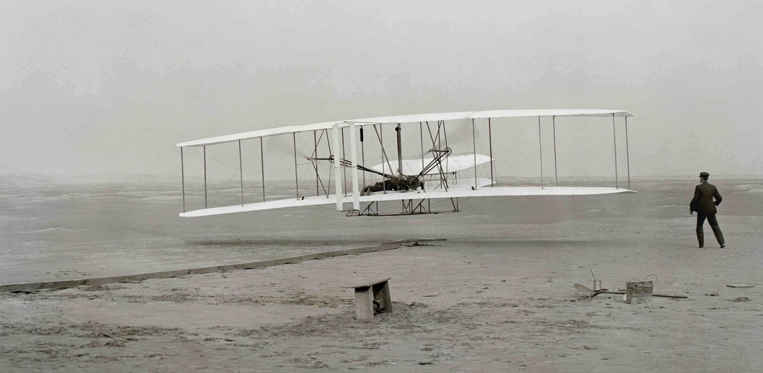 Black and white photograph of a white biplane hovering over a beach. A man in black stands to the right.