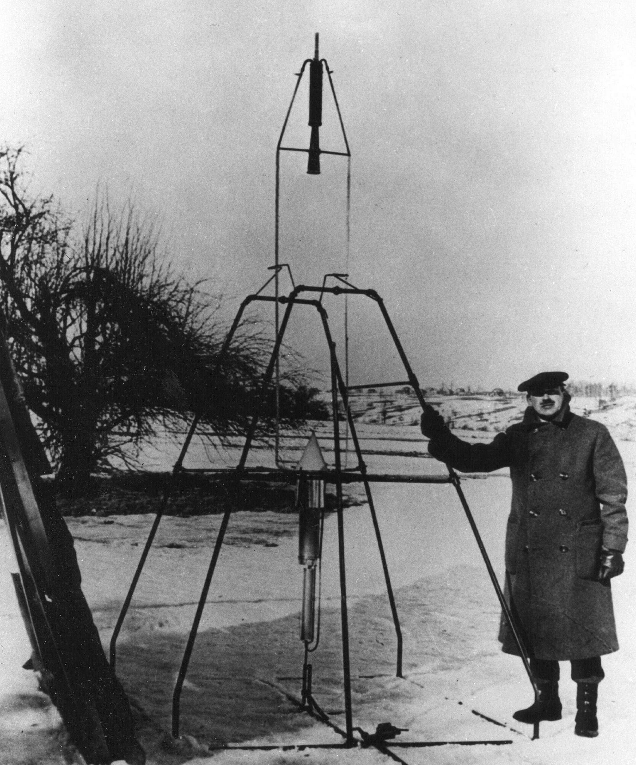A black and white photograph of a man standing next to a launching frame with a small rocket in the center.