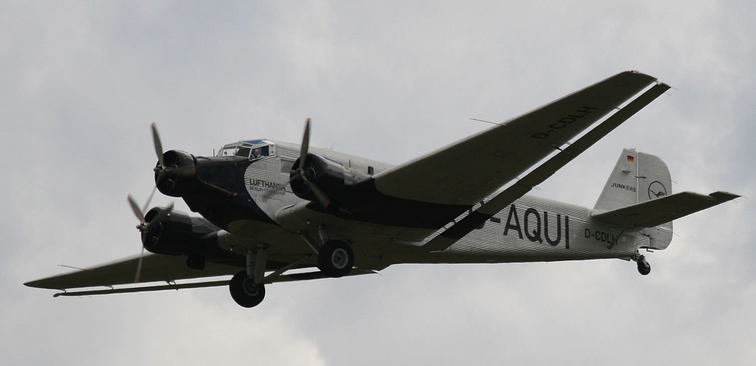 A black and white photograph of an airplane with three propellers and Aqui painted on the fuselage.