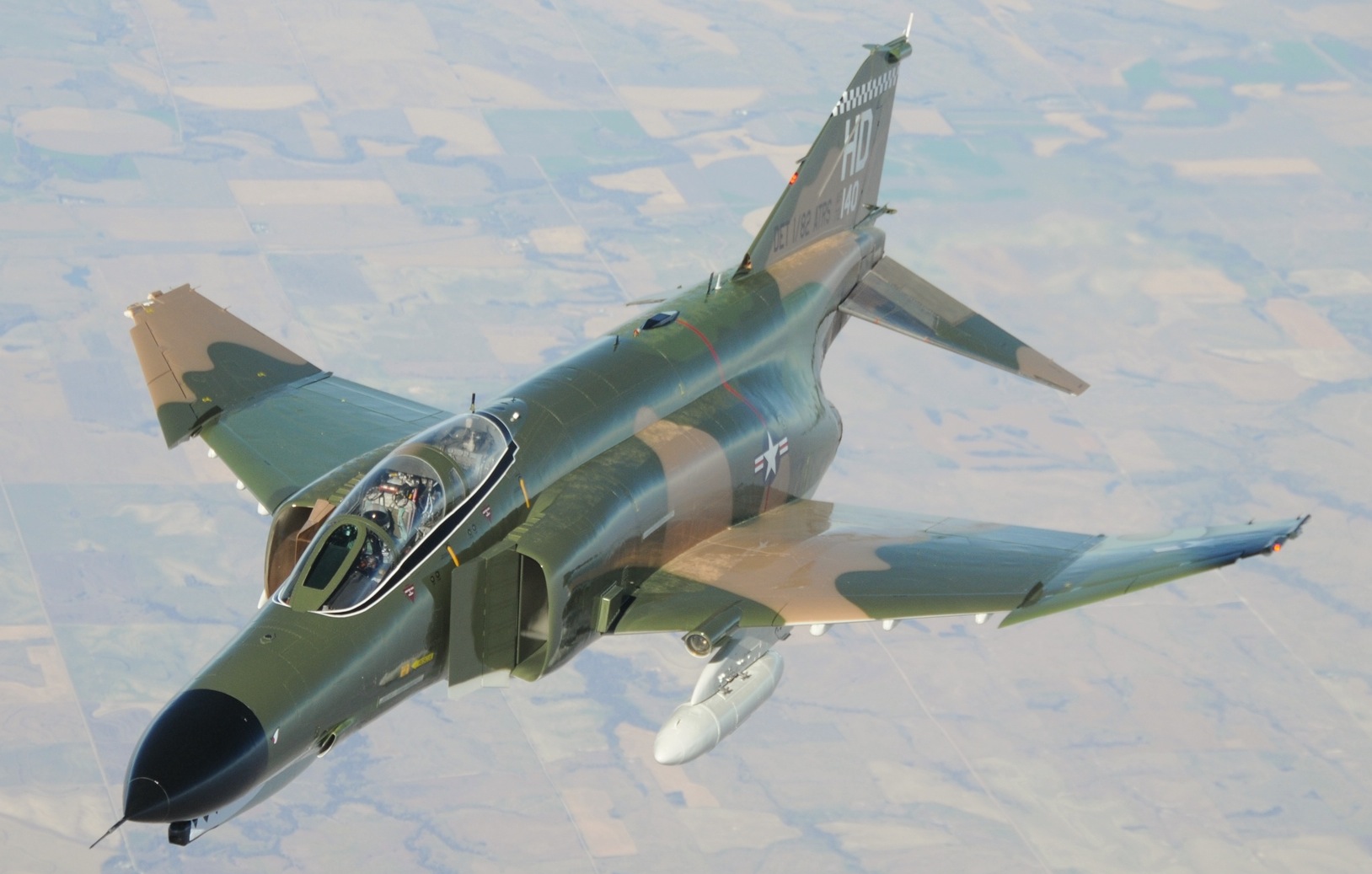 Photograph of a military aircraft painted in a camo design, flying over farmland.