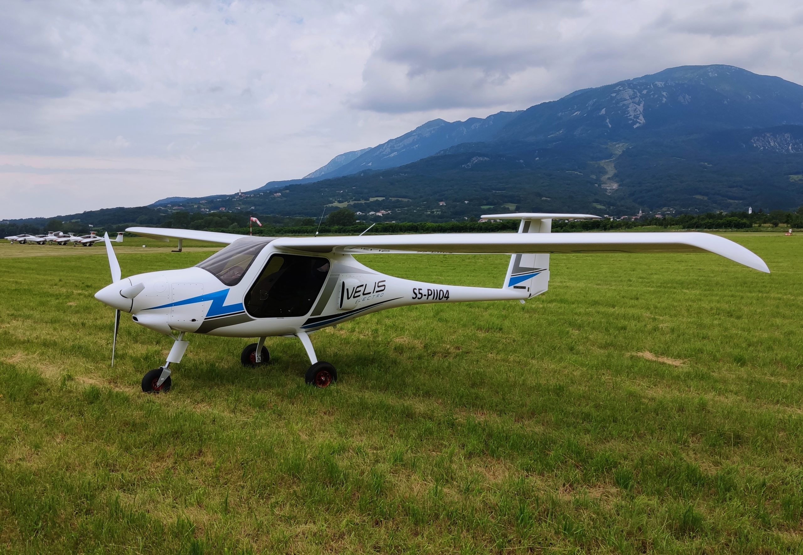 A small white, blue, and gray airplane in a field with a mountain in the background.